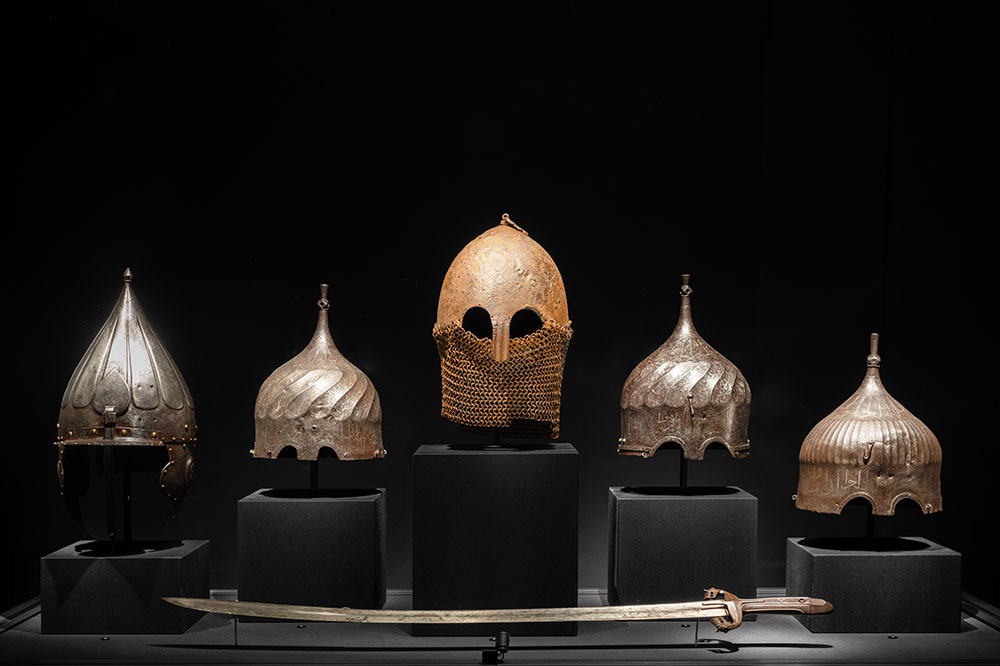 Five helmets and one sword displayed on a museum plinth against a black background. The central Helmet shows an orange iron oxide patina.