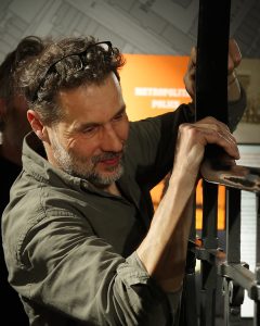 Lead conservator Pavol lifts the plastic coating from the metal handrail. Image shows Pavols head and shoulders, both hands are pushing the plastic rail up from the metalwork which is at head height. 