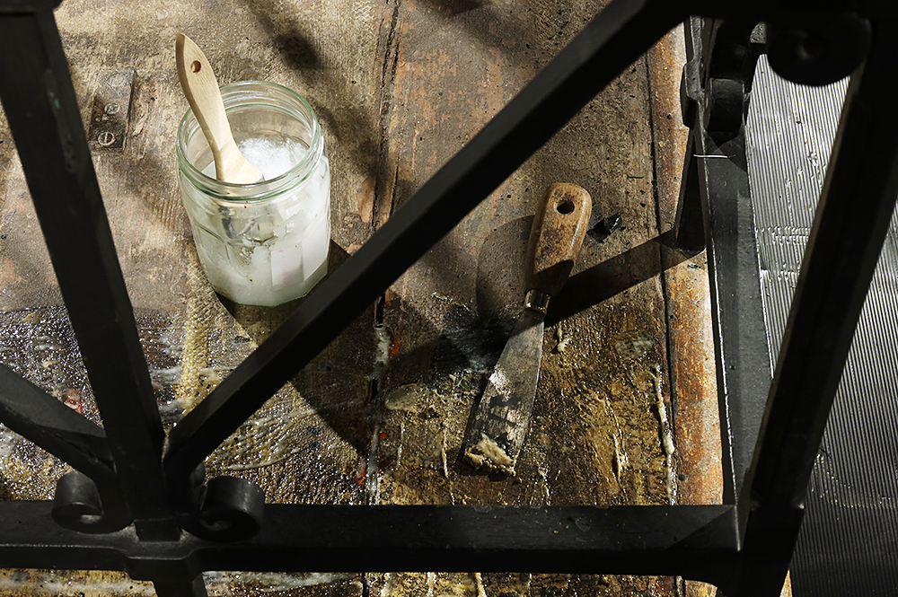 The wooden base of a the dock from Courtroom No.2 at Bow Street Museum of Crime and Justice with a scraper and jar of paste. Some paste has been applied to the surface to soften the adhesive later. The bottom of the image shows a glossier area where the paste has been applied and the top of the image the wood appears dull and matte. The wooden base is seen through a lattice of metal railings.