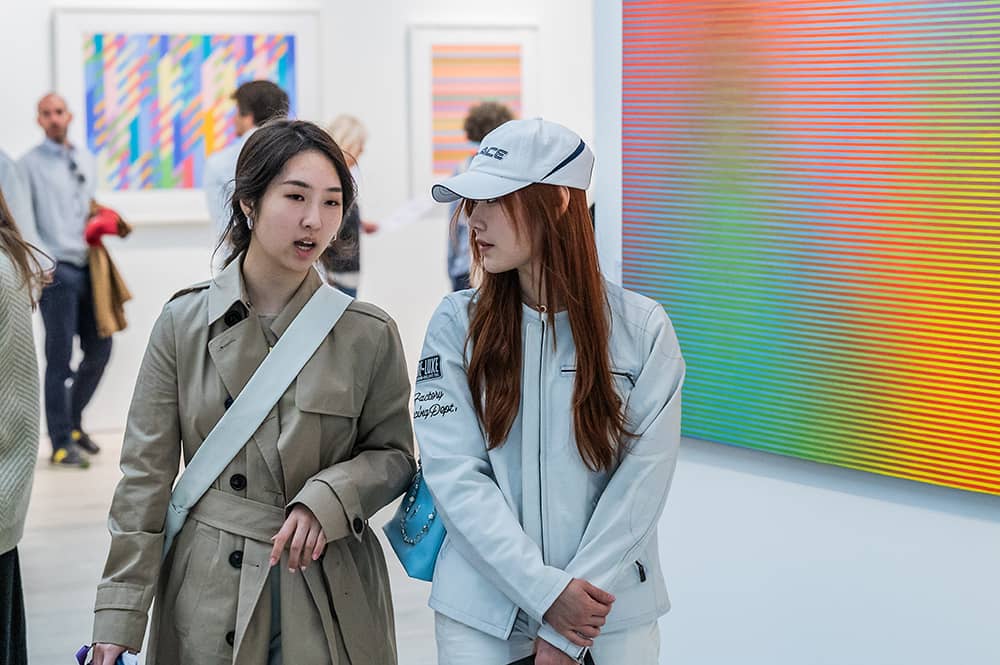 British Art Fair interior, white gallery walls with large colourful artworks. In the foreground two women walk through the fair in conversation.