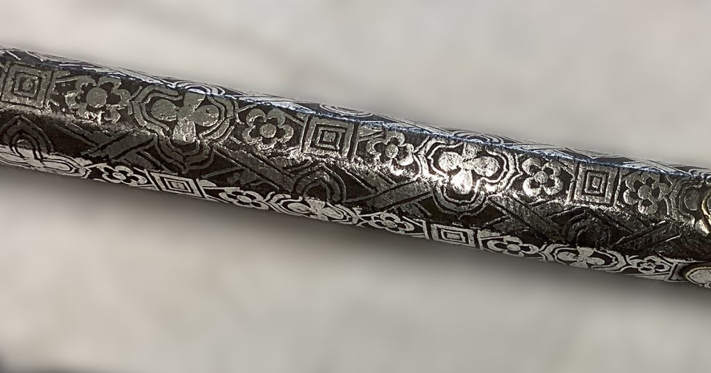 A steel handle that has been cleaned revealing the bright silver inlay against the dark grey iron oxide layer on the steel.