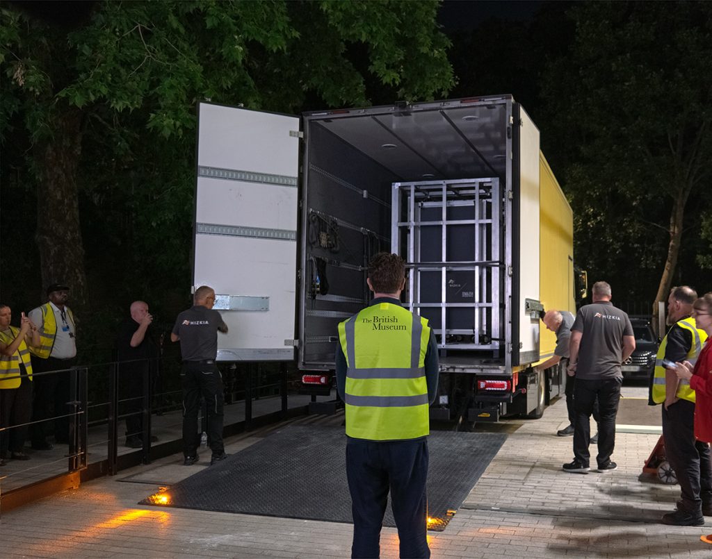 A man in a British Museum high-vis stands in front of the open back of a transport lorry containing a crate. It is night time and around the lorry many people are taking photos.